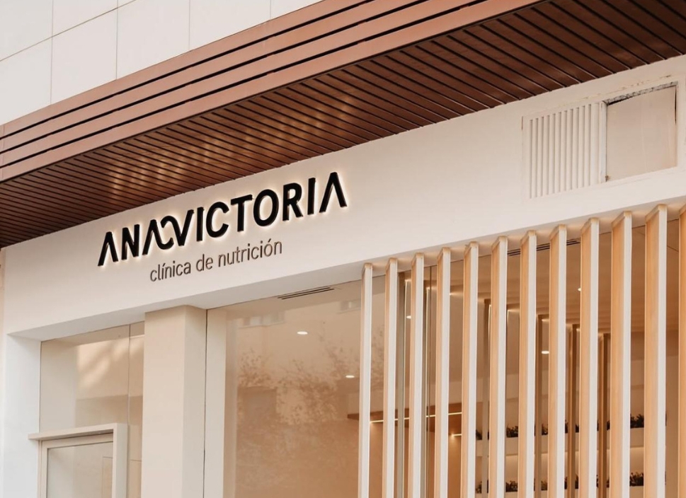 Fotografía del logotipo ANAVICTORIA en la fachada de su clínica de nutrición, en un entorno moderno con acabados en madera y tonos neutros.