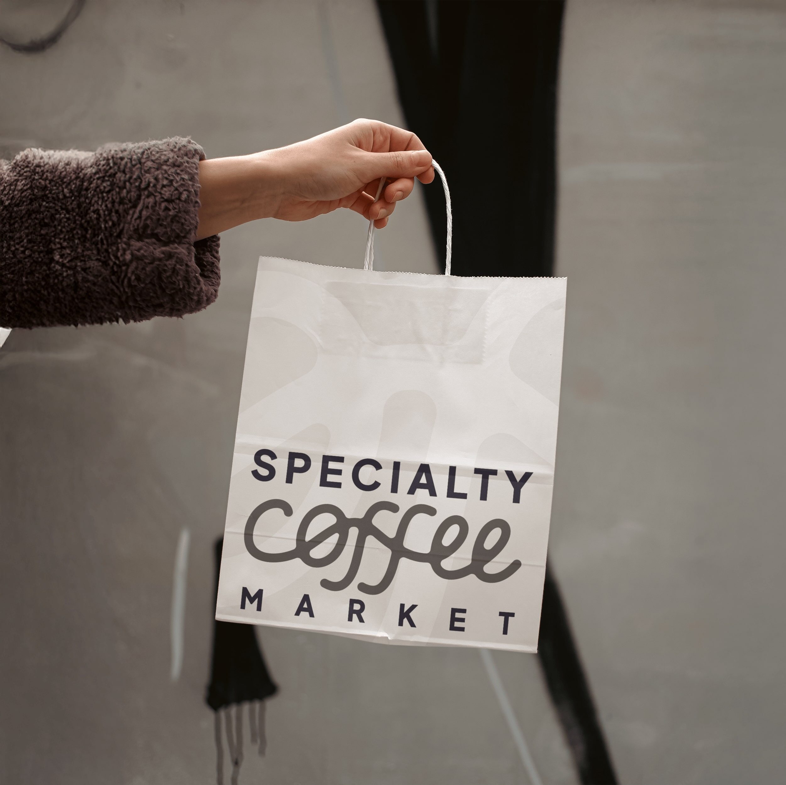 Bolsa de papel blanca con el nuevo logotipo de Specialty Coffee Market impreso, sostenida por una mano. Muestra la aplicación del branding en packaging físico y su legibilidad en contextos reales.