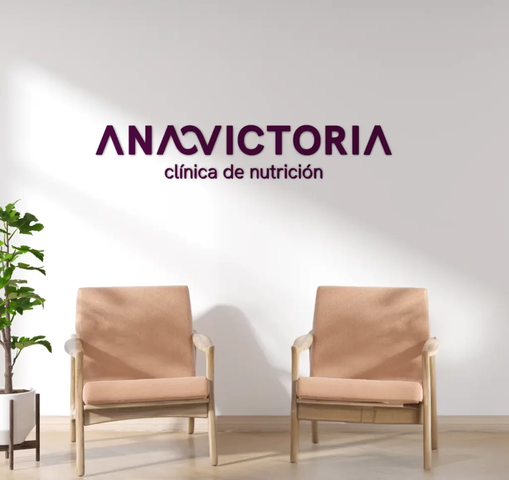 Render del logotipo ANAVICTORIA clínica de nutrición en una pared blanca de una sala de espera minimalista con sillas de madera clara.